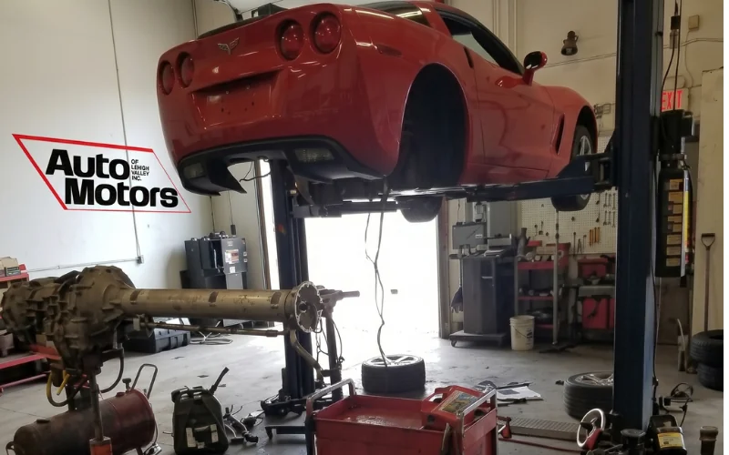 A red car raised on a lift in a repair shop, with a removed drivetrain assembly on the floor beneath it.